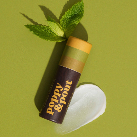 A large lip balm tube with a green cap and gold accents labeled 'Poppy & Pout', displayed next to a pool of balm and a mint leaf on a green background.