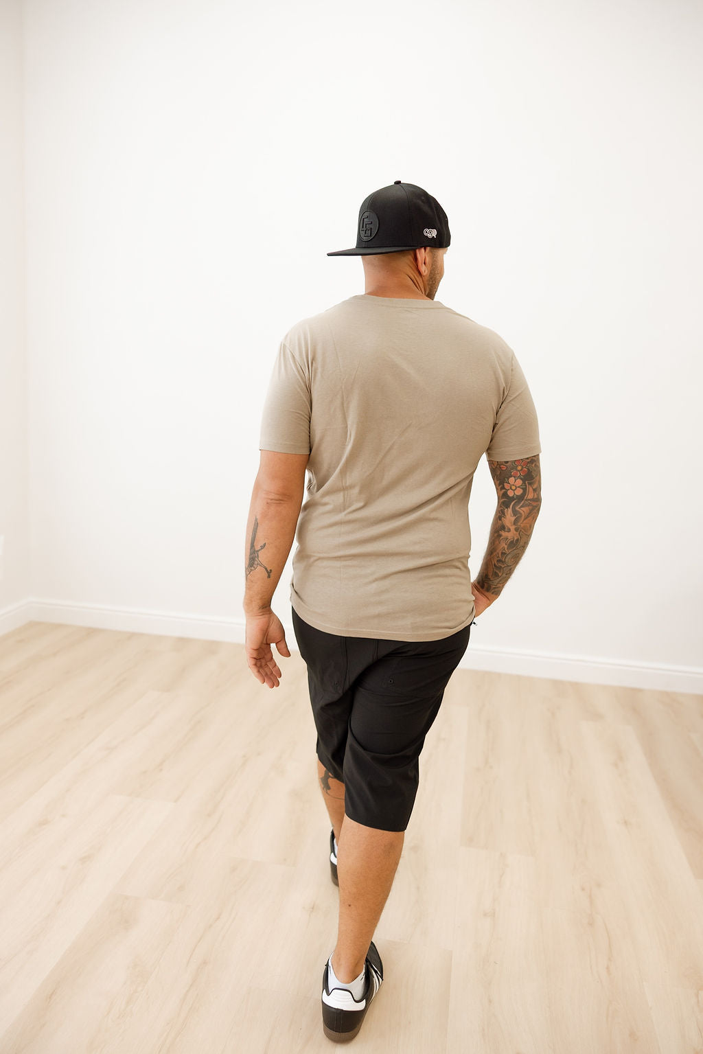 Man wearing a beige t-shirt and black shorts walking in a room with a white wall and wooden floor.