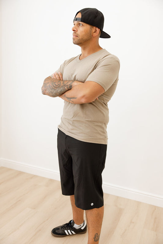 Man wearing a beige t-shirt, black shorts, and a black cap standing against a white wall.