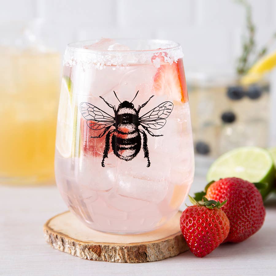 Stemless wine glass with a bee design on a wooden coaster, surrounded by strawberries and limes.