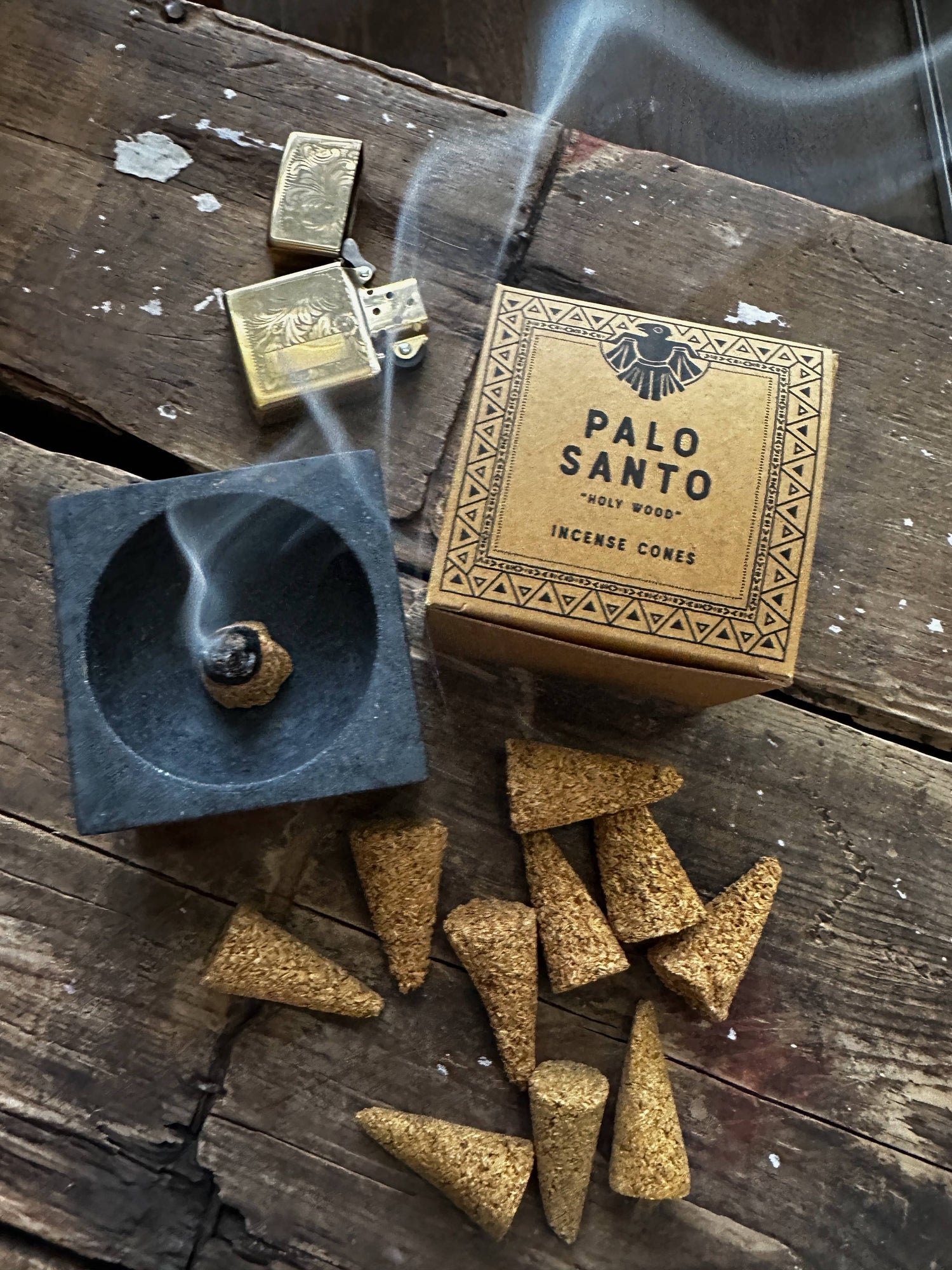 A box of Palo Santo incense cones displayed alongside some cones that are burning on a black holder, emitting smoke, placed on a rustic wooden surface.