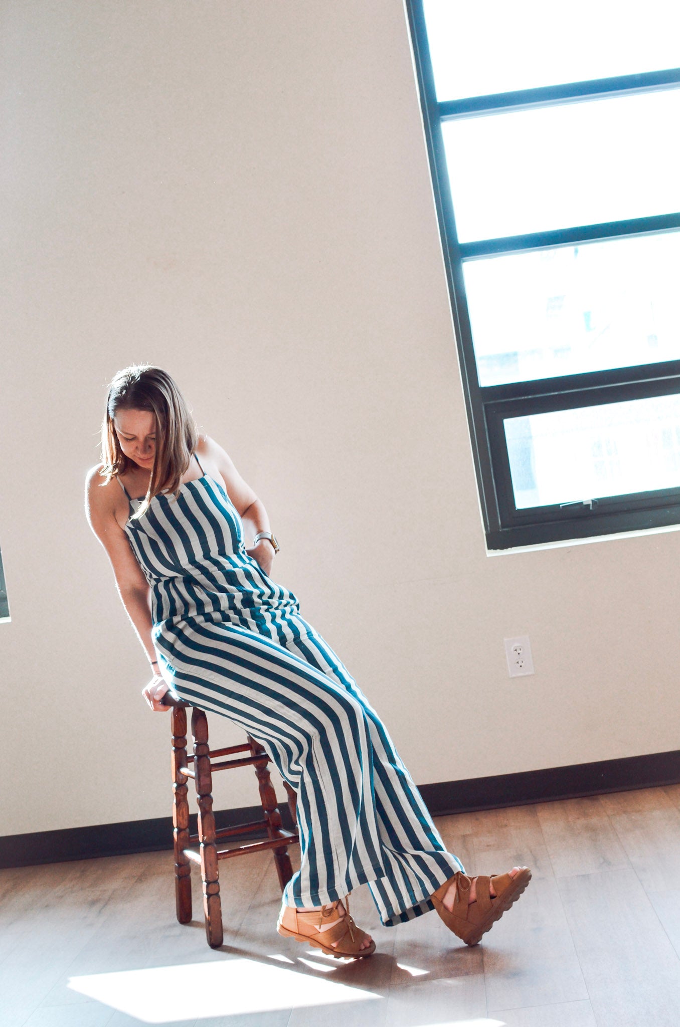 Woman in a striped jumpsuit sitting on a wooden stool in a room with large windows.