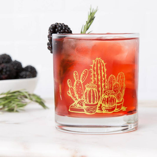 Glass of red cocktail with cactus design, blackberries, and rosemary on a white background