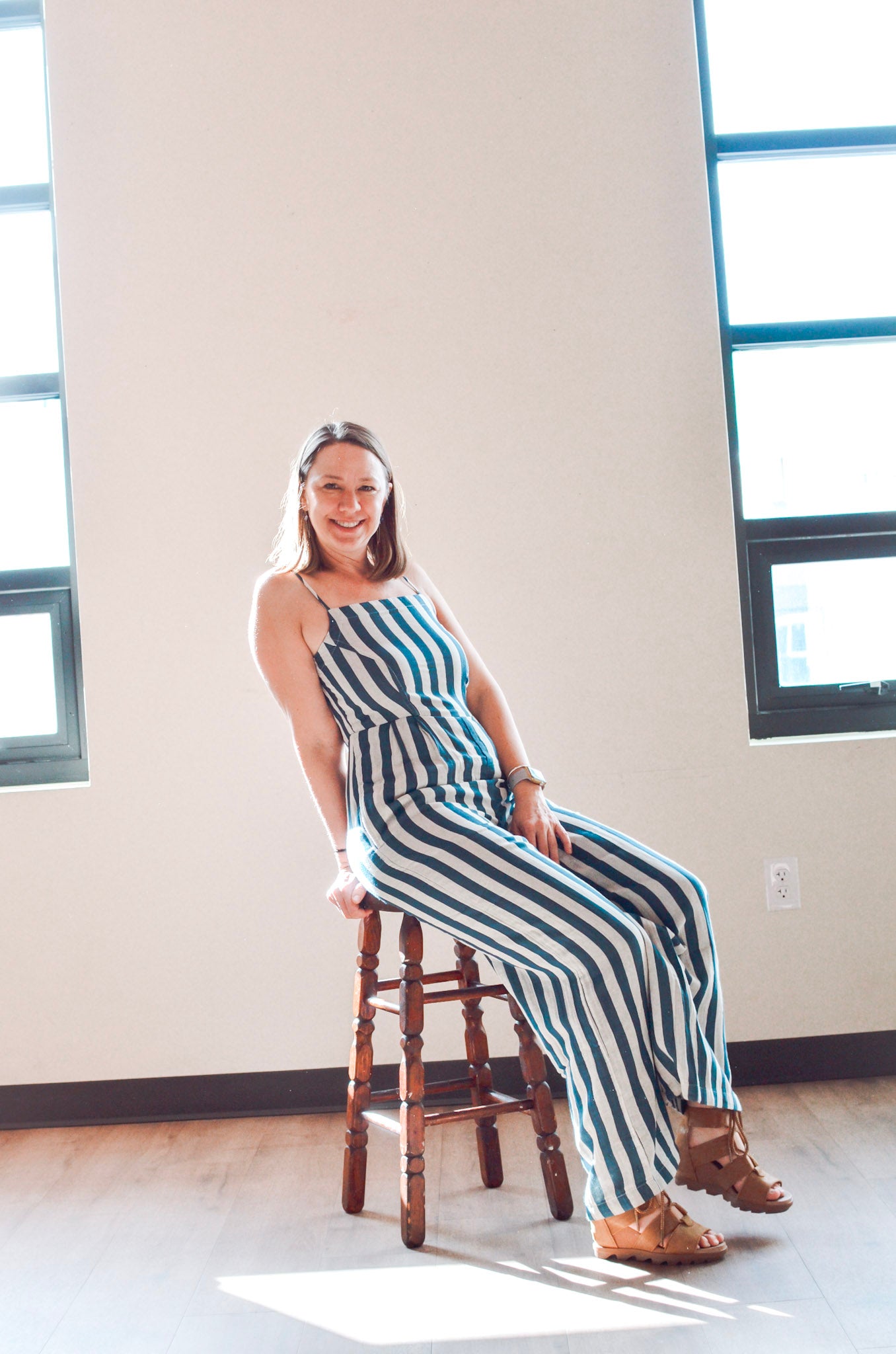 Woman in a striped dress sitting on a wooden stool in a room with large windows.