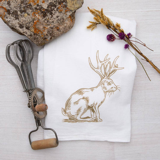 White towel with a gold hare print, whisk, and stone on a light background