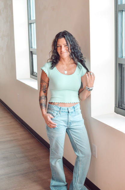 Woman wearing a light green top and blue jeans standing in a room with large windows.