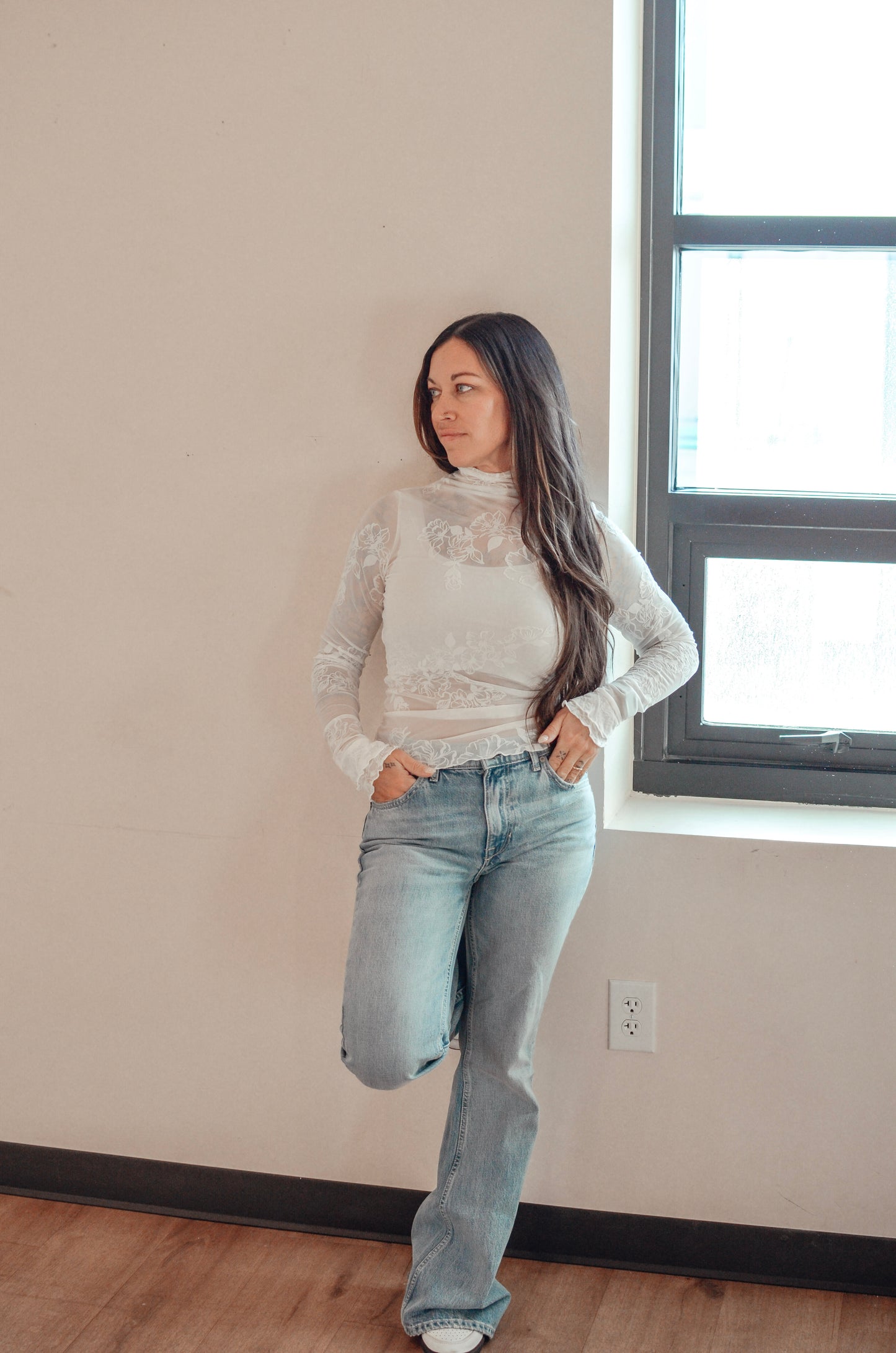 Woman wearing a white lace top and blue jeans standing in a room with a window.