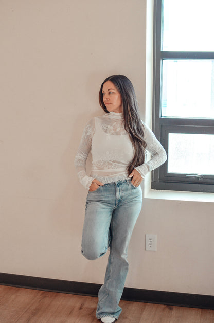 Woman wearing a white lace top and blue jeans standing in a room with a window.