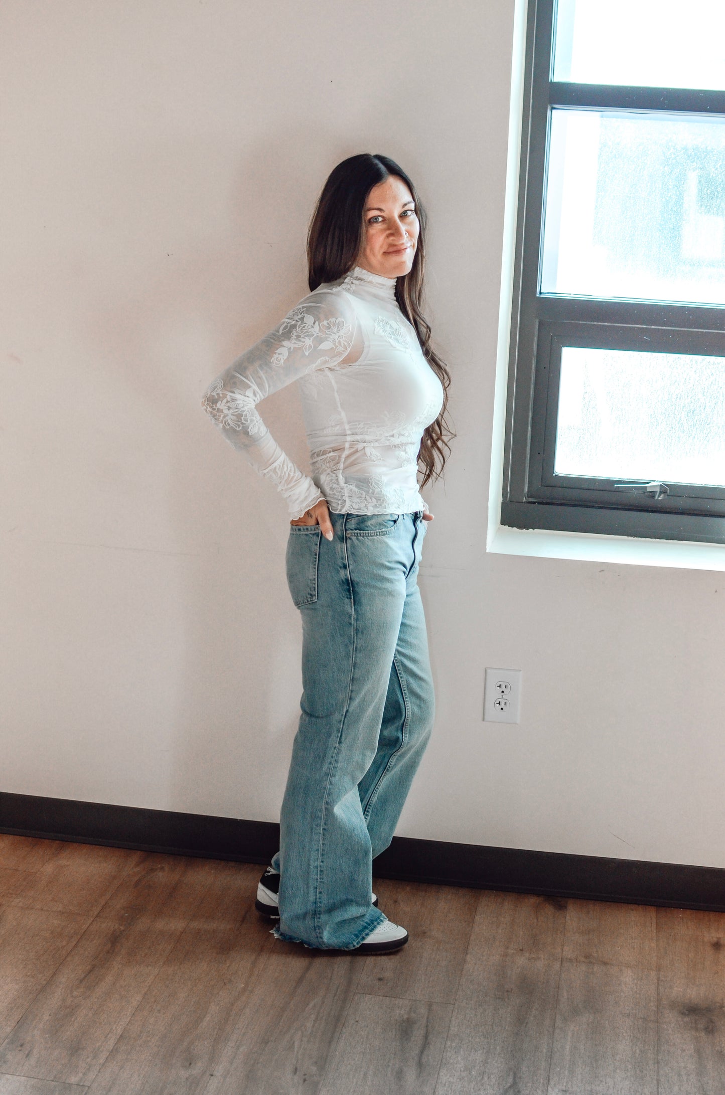 Woman wearing a white long-sleeve top and light blue jeans standing in a room with a window.