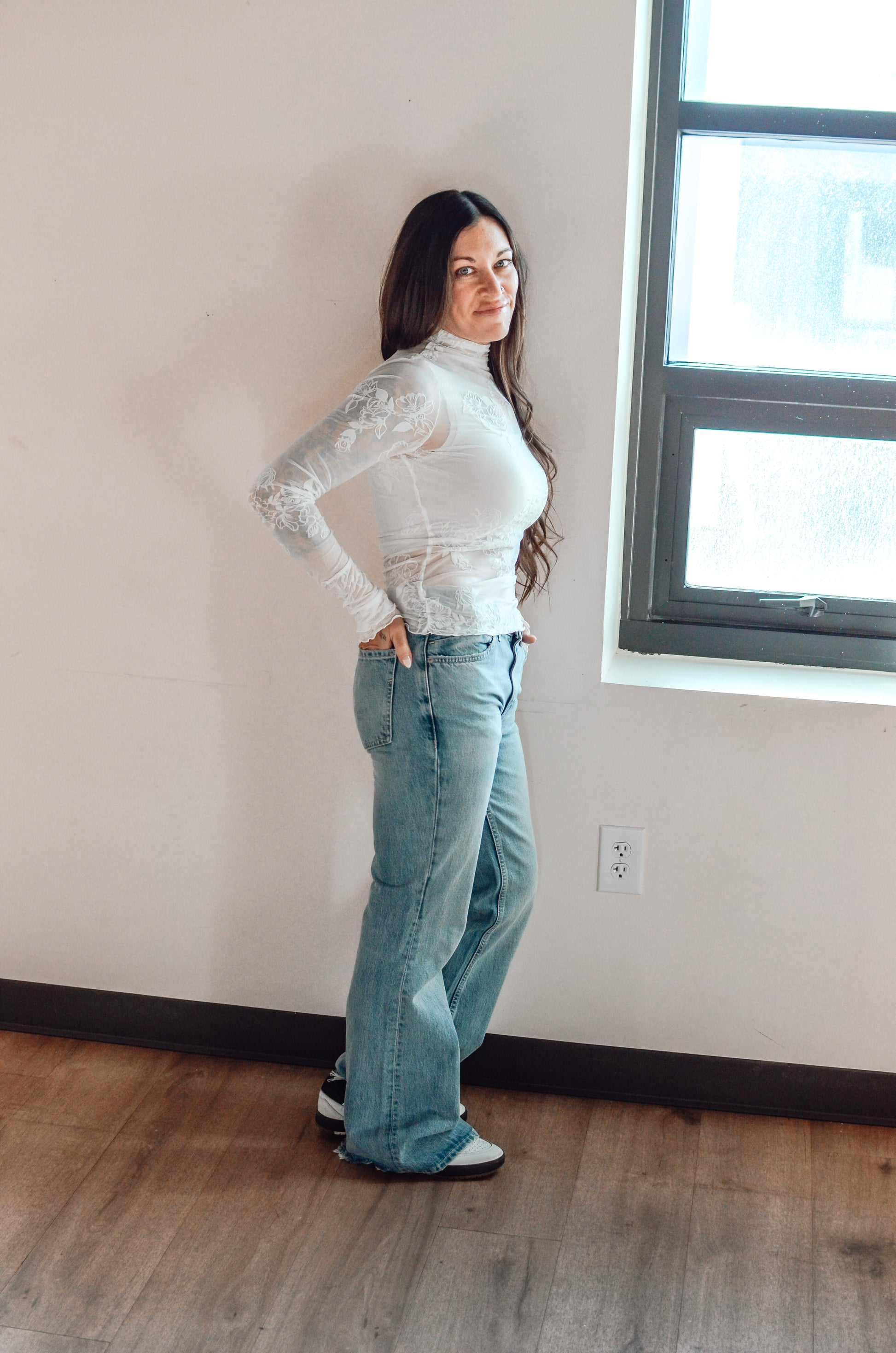 Woman wearing a white long-sleeve top and light blue jeans standing in a room with a window.