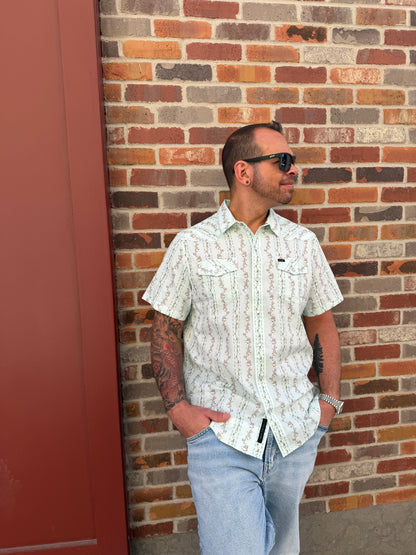 Person wearing a white patterned shirt and blue jeans against a brick wall.
