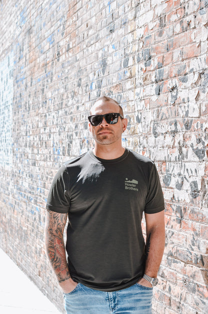 Man wearing a black t-shirt with a logo standing against a brick wall.