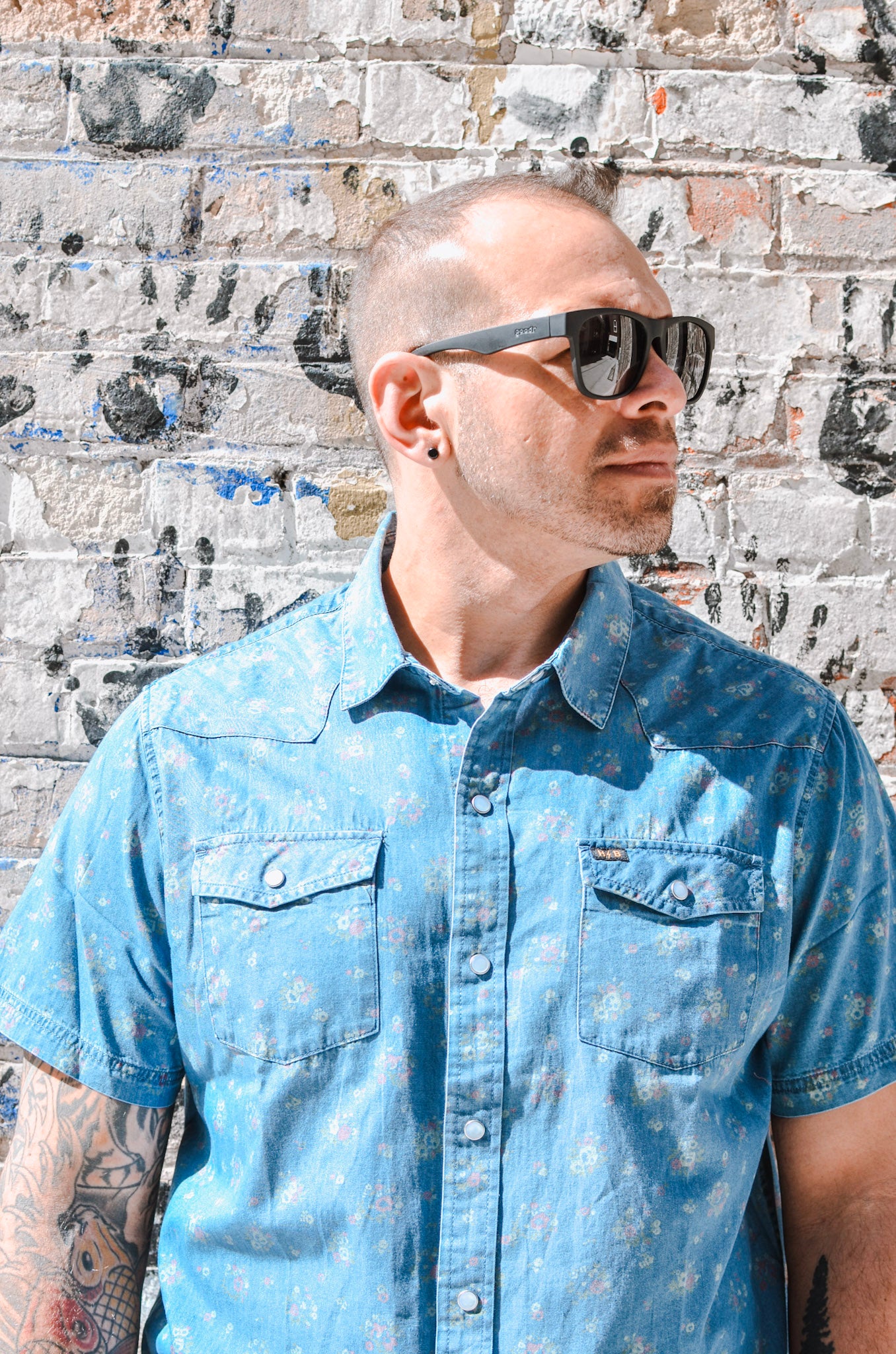 Man wearing sunglasses and a blue shirt against a textured wall.