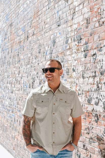 Man wearing sunglasses and a beige shirt standing against a textured brick wall.