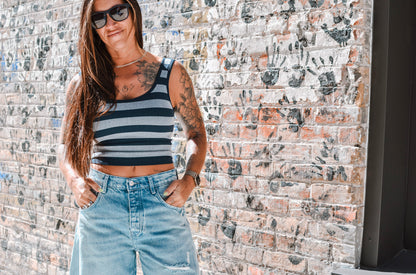 Woman wearing a striped tank top and denim shorts standing against a textured wall.