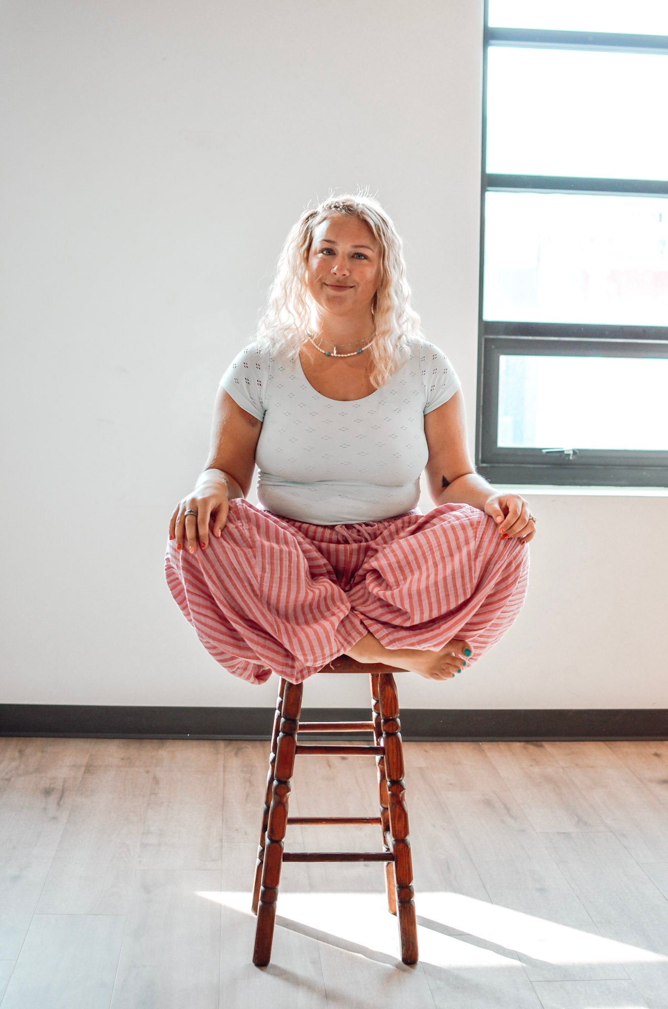 Woman sitting on a wooden stool wearing pink striped pants and a white top in a bright room.