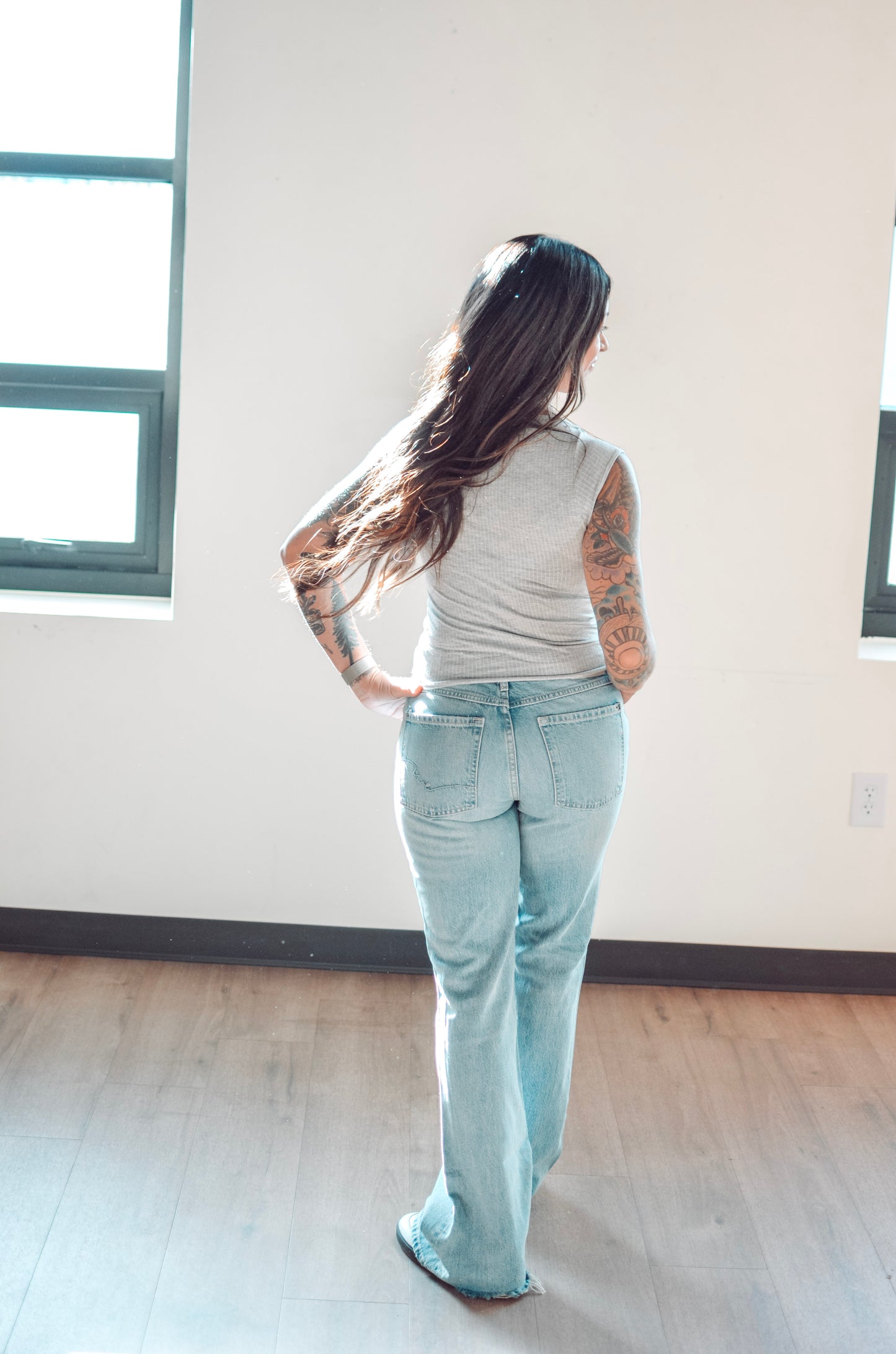 Person wearing light blue jeans and a gray top standing in a room with large windows.
