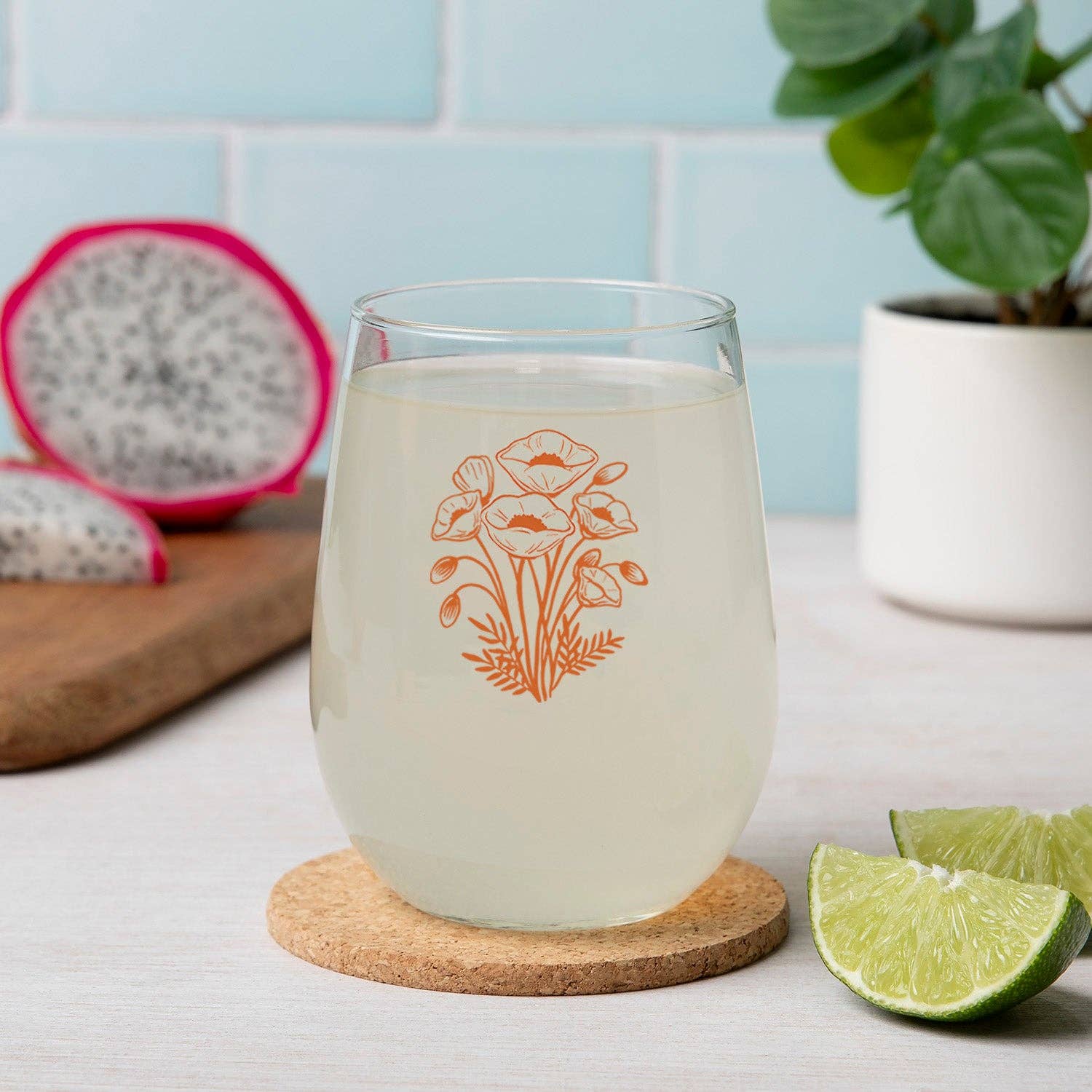 Clear glass with floral design on a cork coaster, dragon fruit, lime, and plant in the background.
