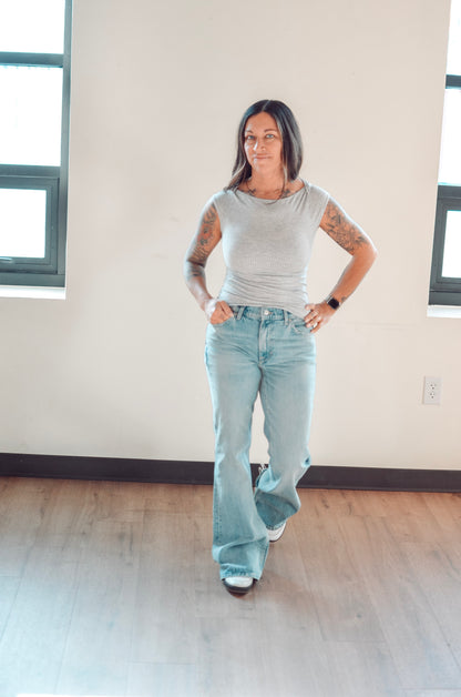 Woman wearing a gray sleeveless top and light blue jeans standing in a room with large windows.