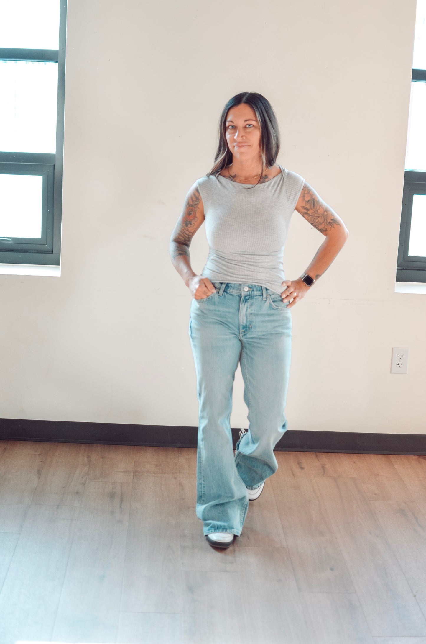 Woman wearing a gray sleeveless top and light blue jeans standing in a room with large windows.
