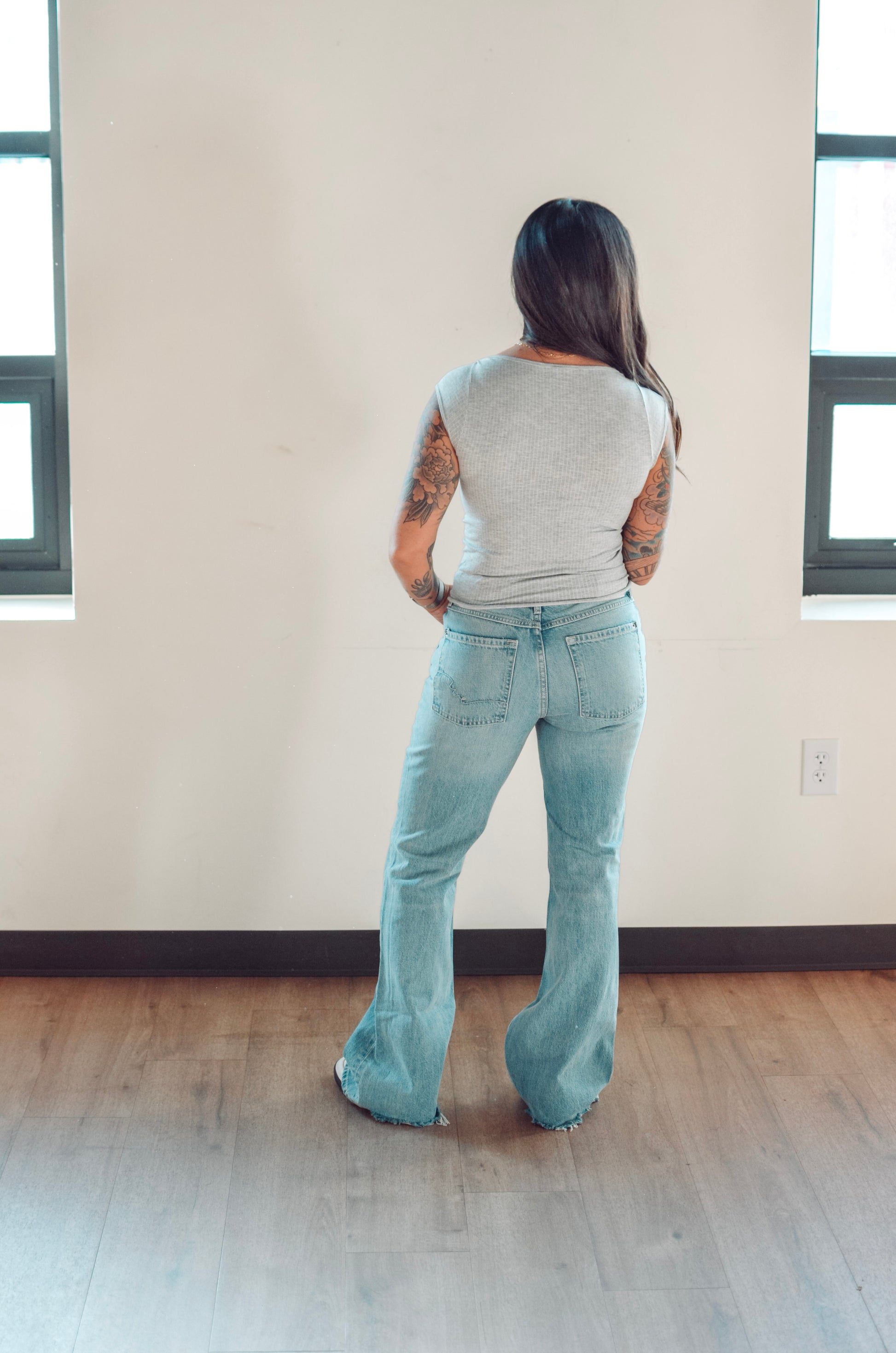 Person wearing light blue jeans and a gray top standing in a room with wooden flooring and a white wall.