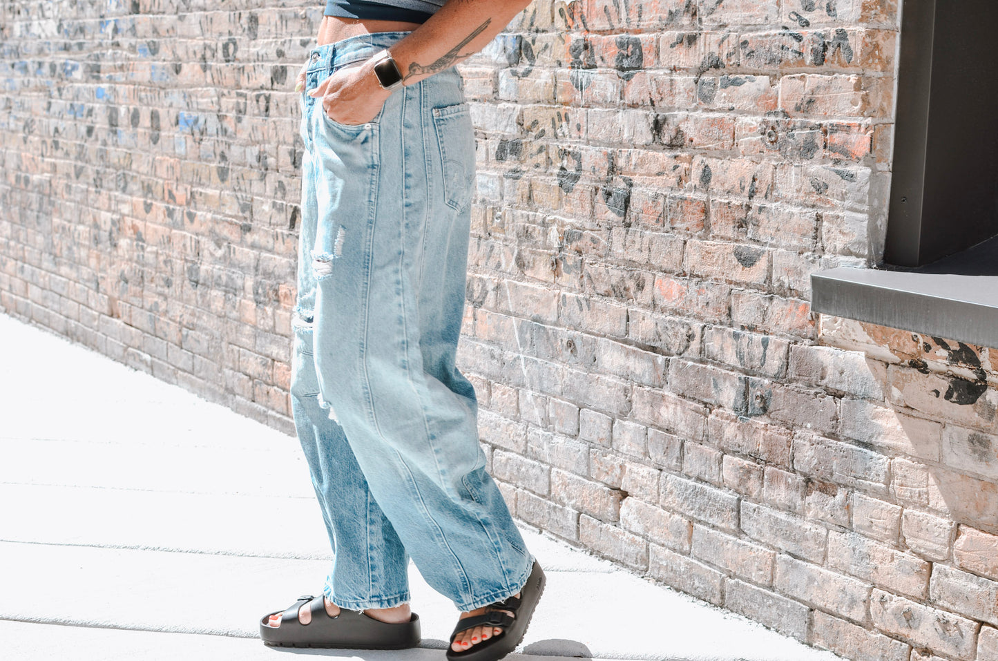 Person wearing light blue jeans and black sandals standing against a stone wall.