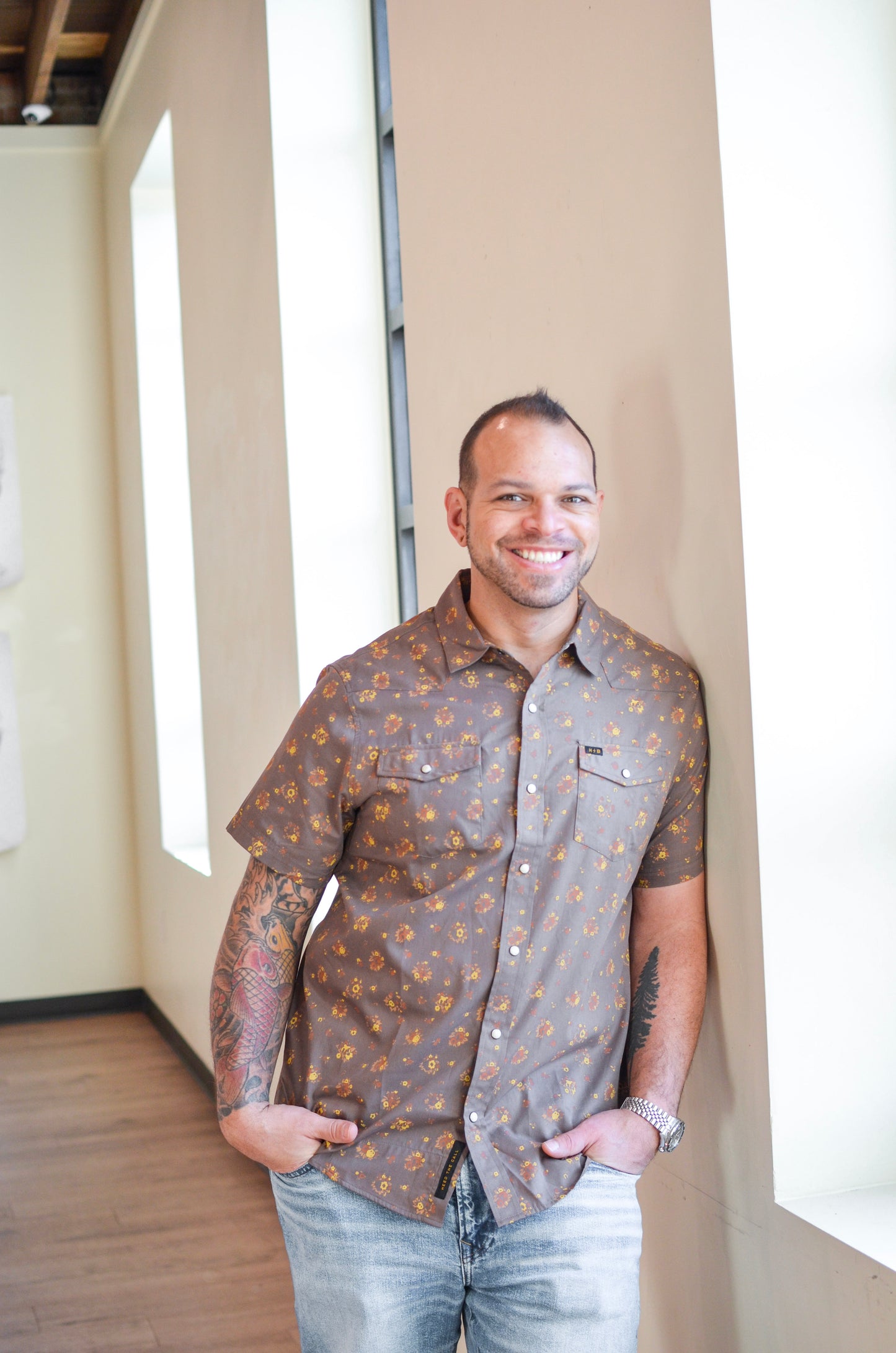Man wearing a patterned shirt and jeans standing in a room with large windows.