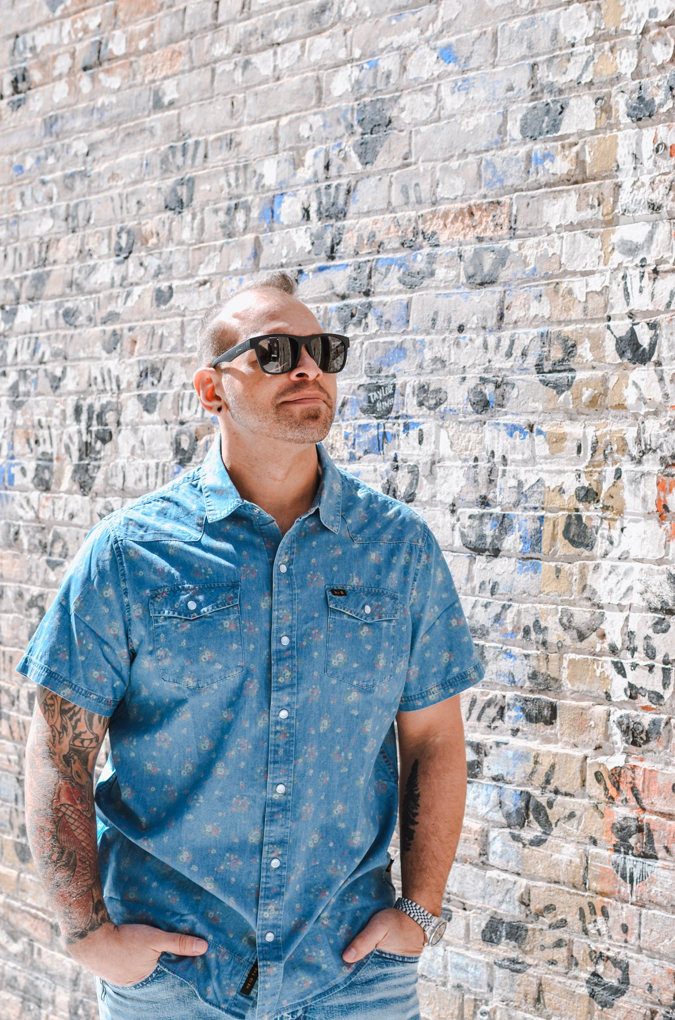 Man wearing sunglasses and a blue shirt against a textured brick wall