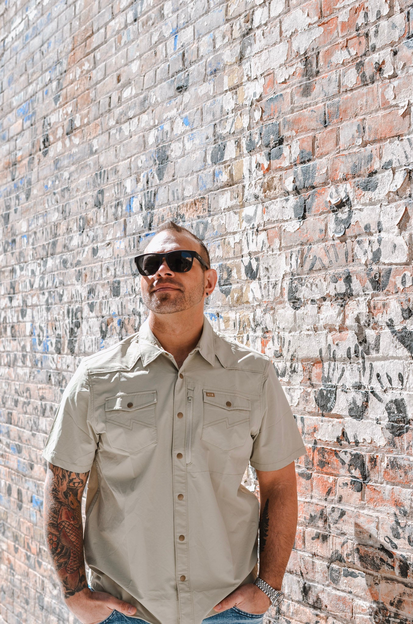 Man wearing sunglasses and a beige shirt against a brick wall