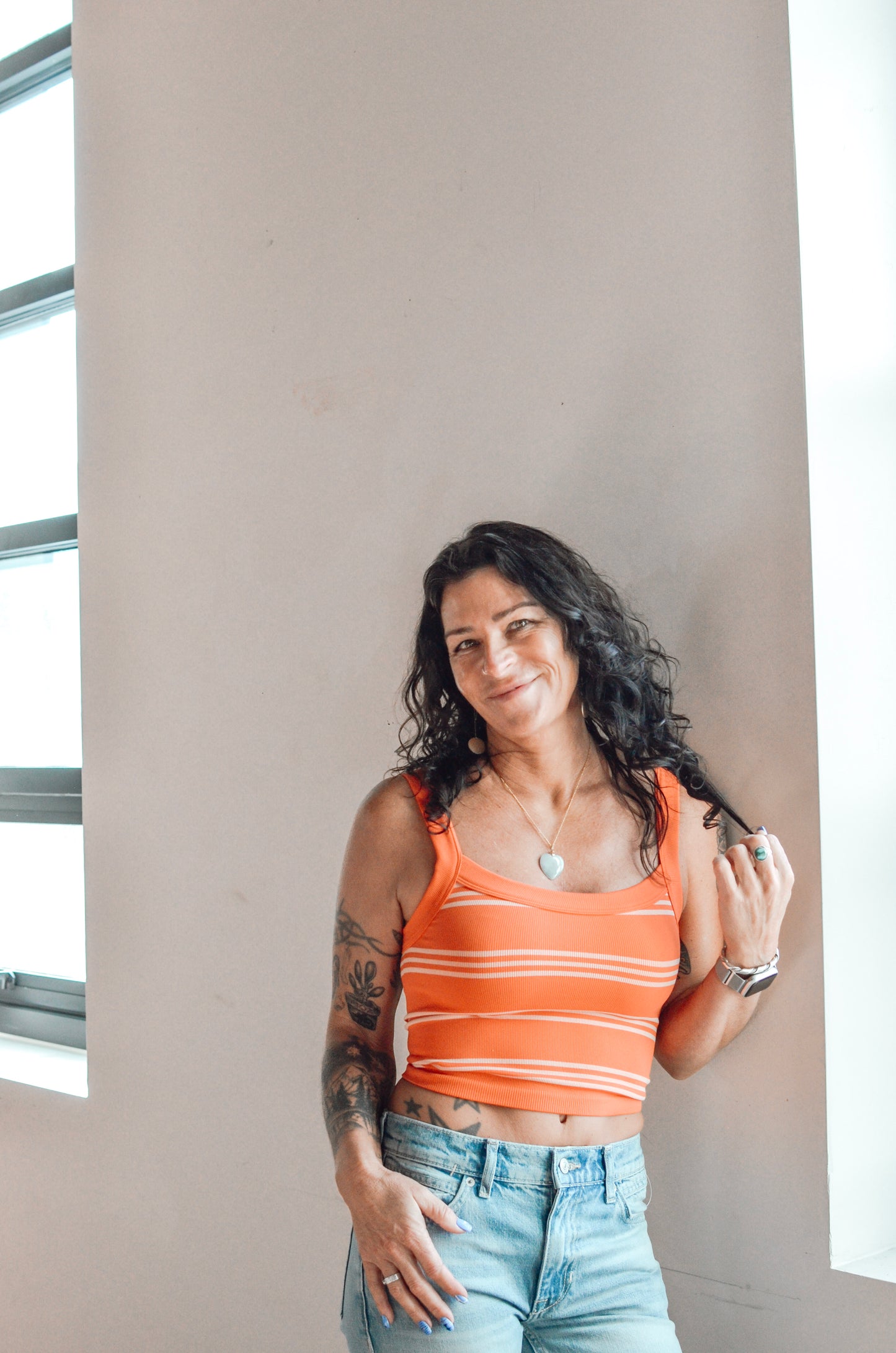 Woman wearing an orange striped tank top and blue jeans standing against a plain wall.