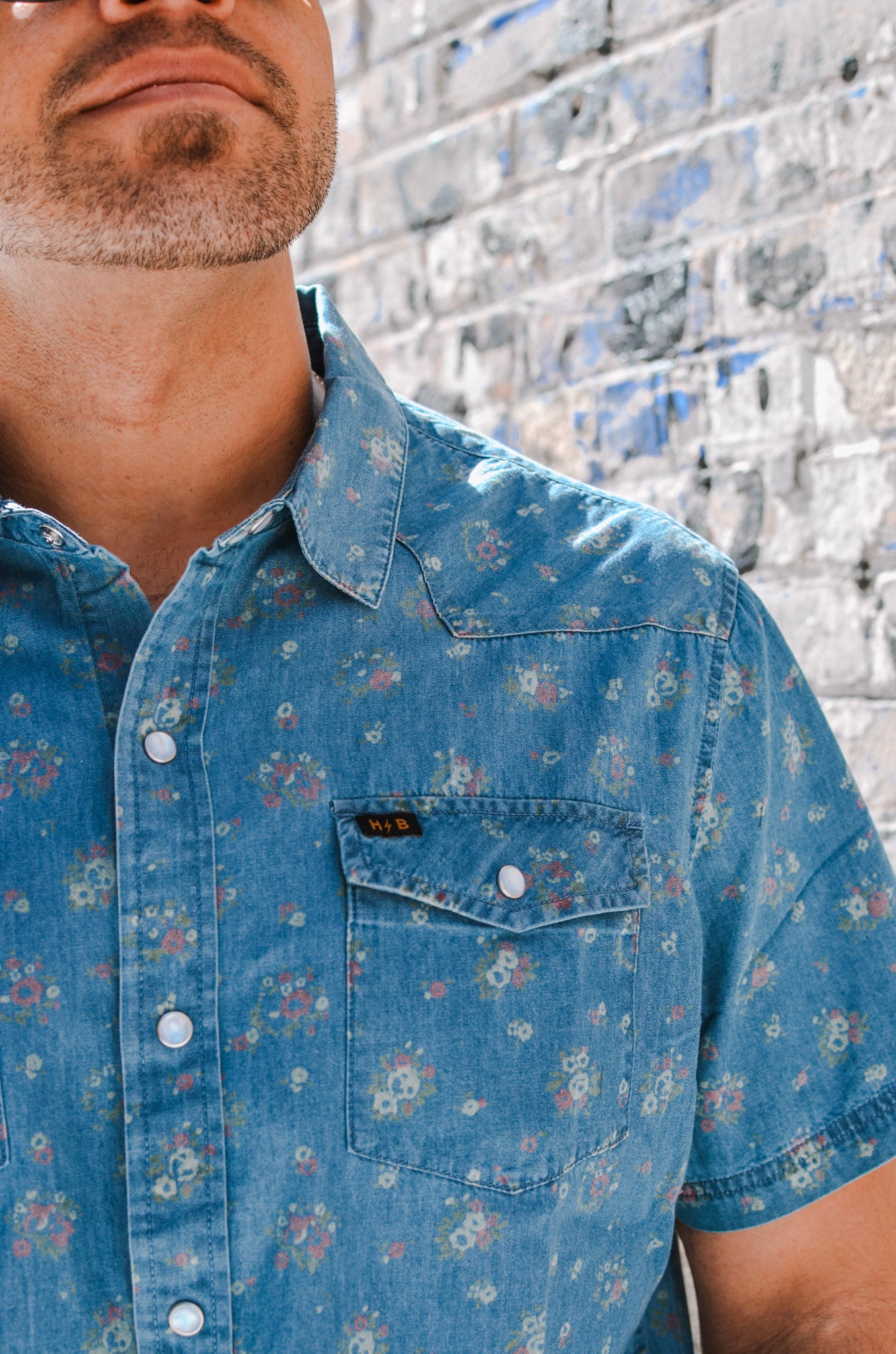 Man wearing a blue floral short-sleeve shirt against a textured wall.