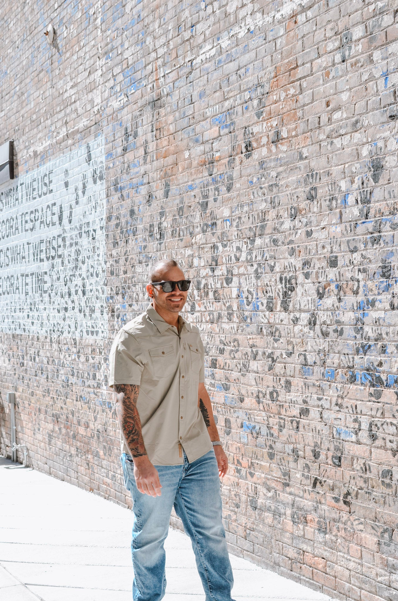 Man standing against a textured brick wall wearing sunglasses and a beige shirt.