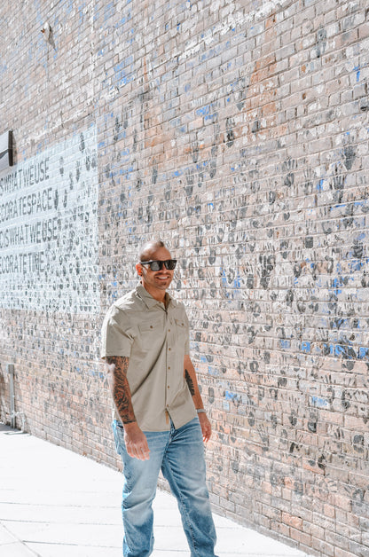 Man standing against a textured brick wall wearing sunglasses and a beige shirt.