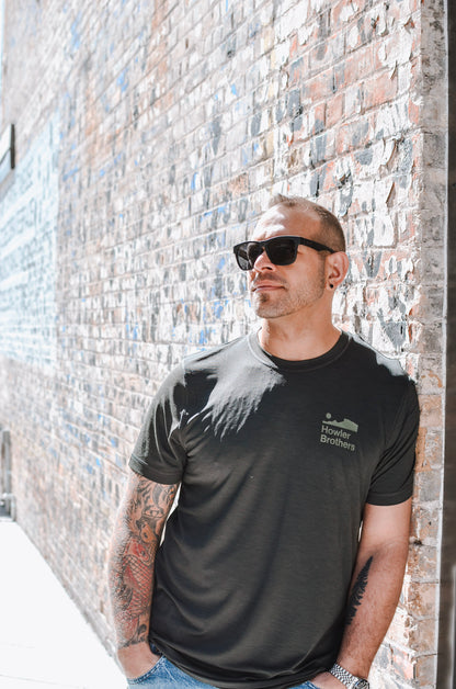 Man wearing a black t-shirt with a logo against a brick wall.