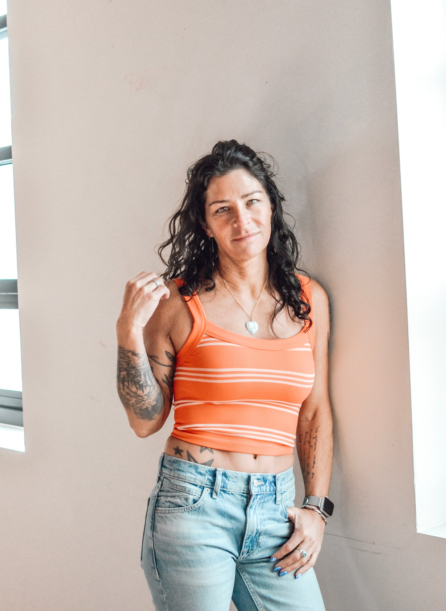 Woman wearing an orange striped tank top and light blue jeans against a neutral wall.