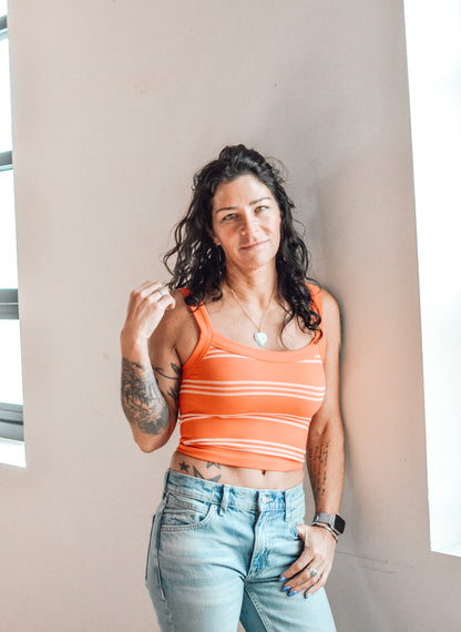 Woman wearing an orange striped tank top and light blue jeans against a neutral wall.