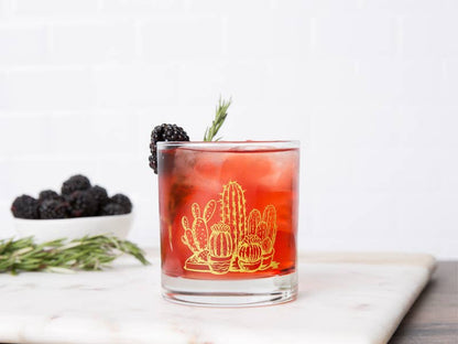 Cocktail in a glass with cactus design, blackberries, and rosemary on a marble surface.