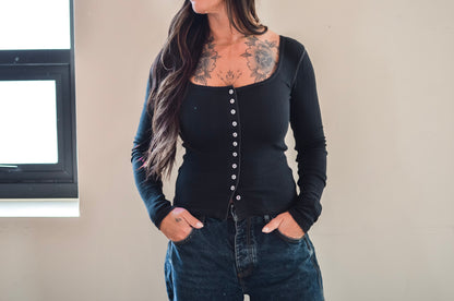 Person wearing a black long-sleeve top with buttons and blue jeans against a neutral background