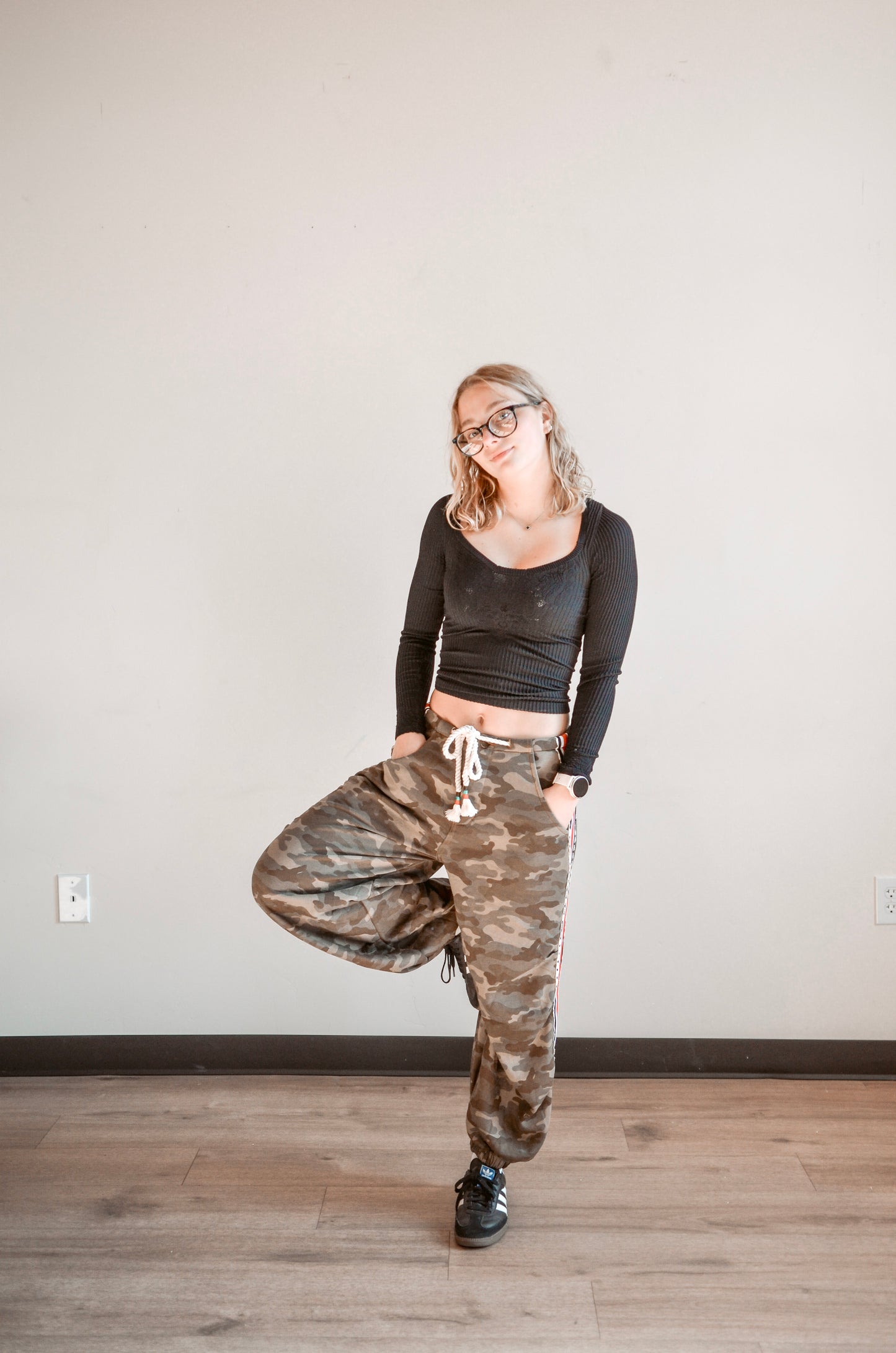Person wearing a black long-sleeve top and camouflage pants standing on a wooden floor with a white wall background.