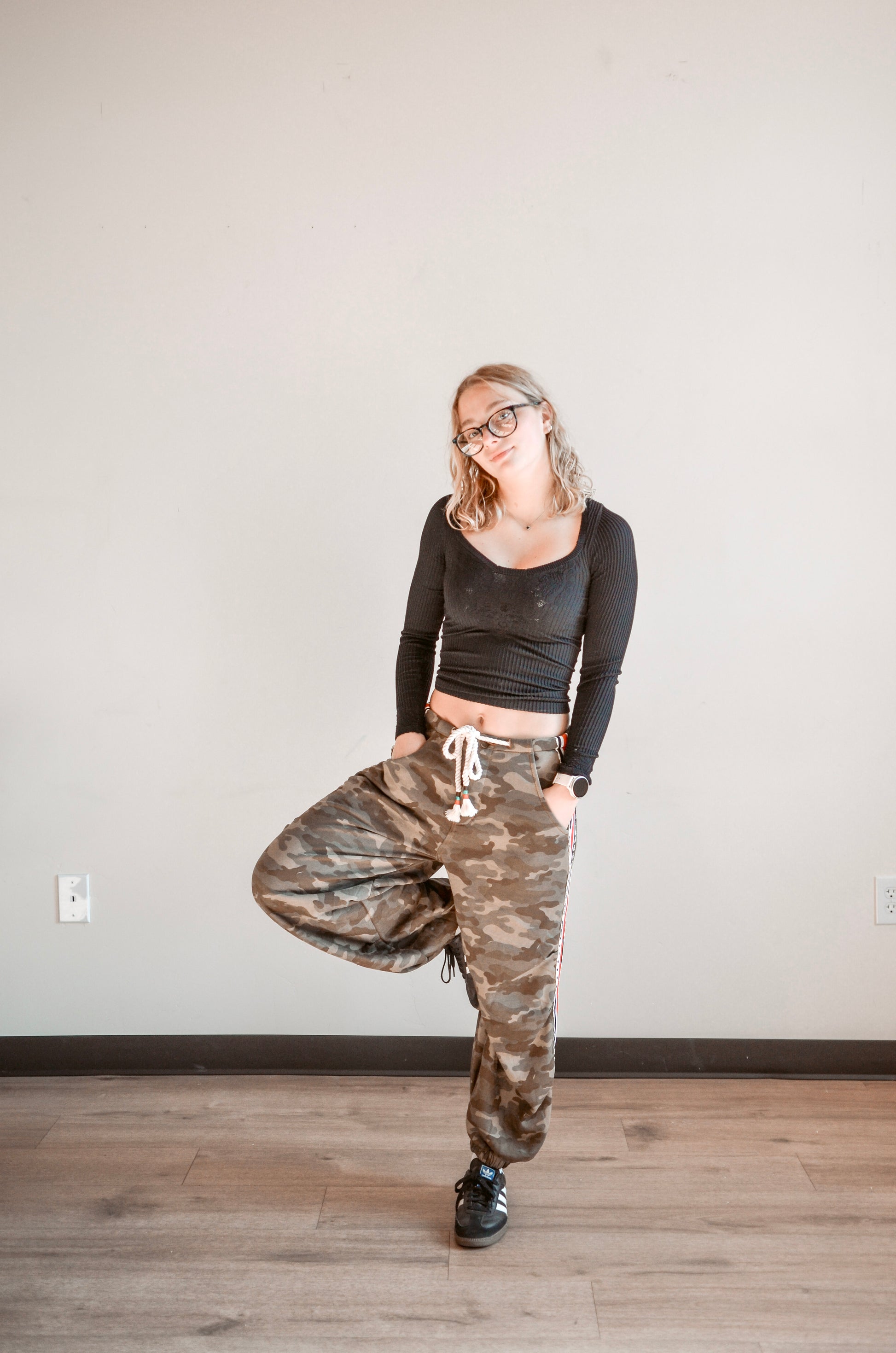 Person wearing a black long-sleeve top and camouflage pants standing on a wooden floor with a white wall background.