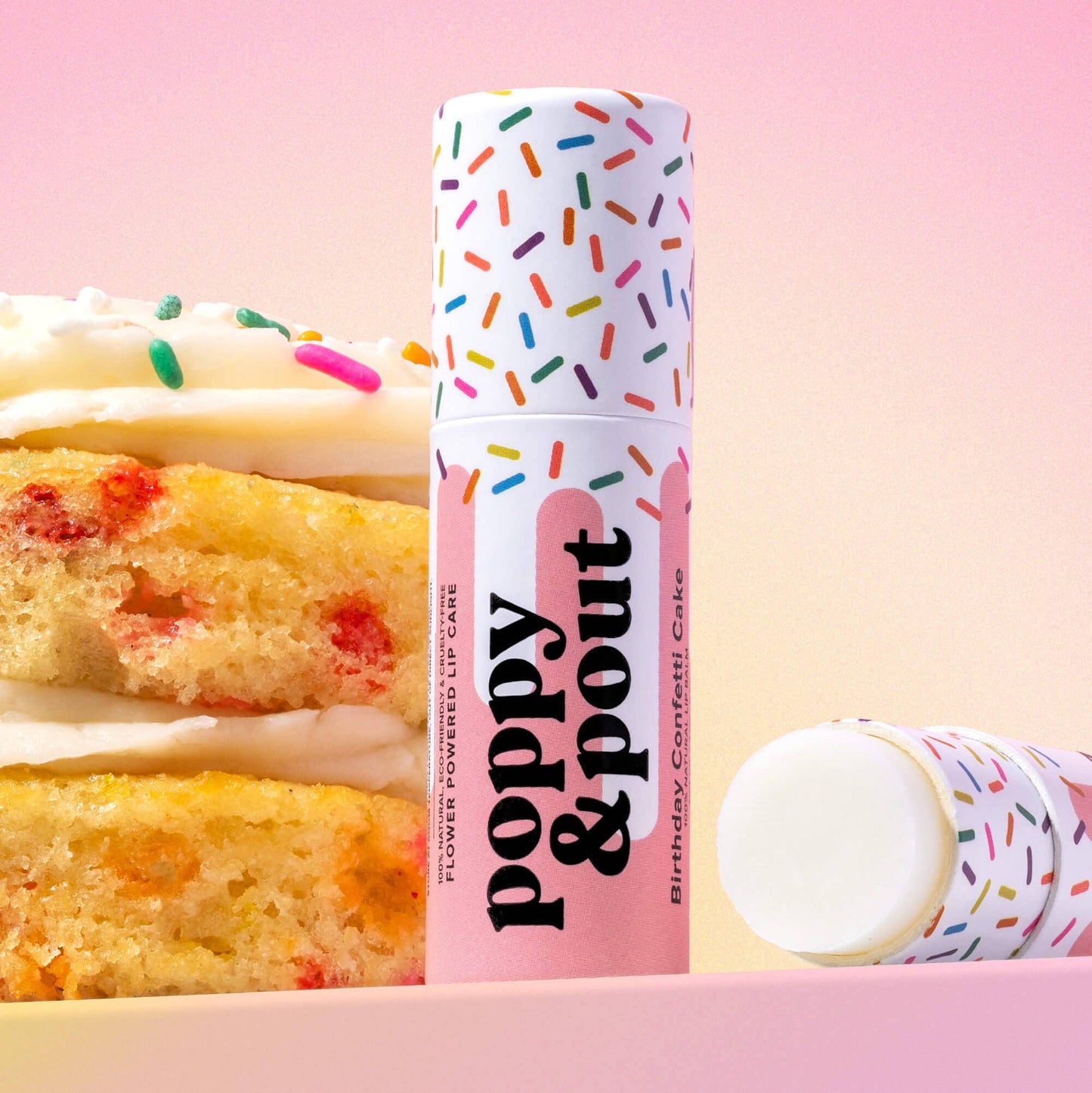 Lip balm with colorful packaging next to a cake on a pink background
