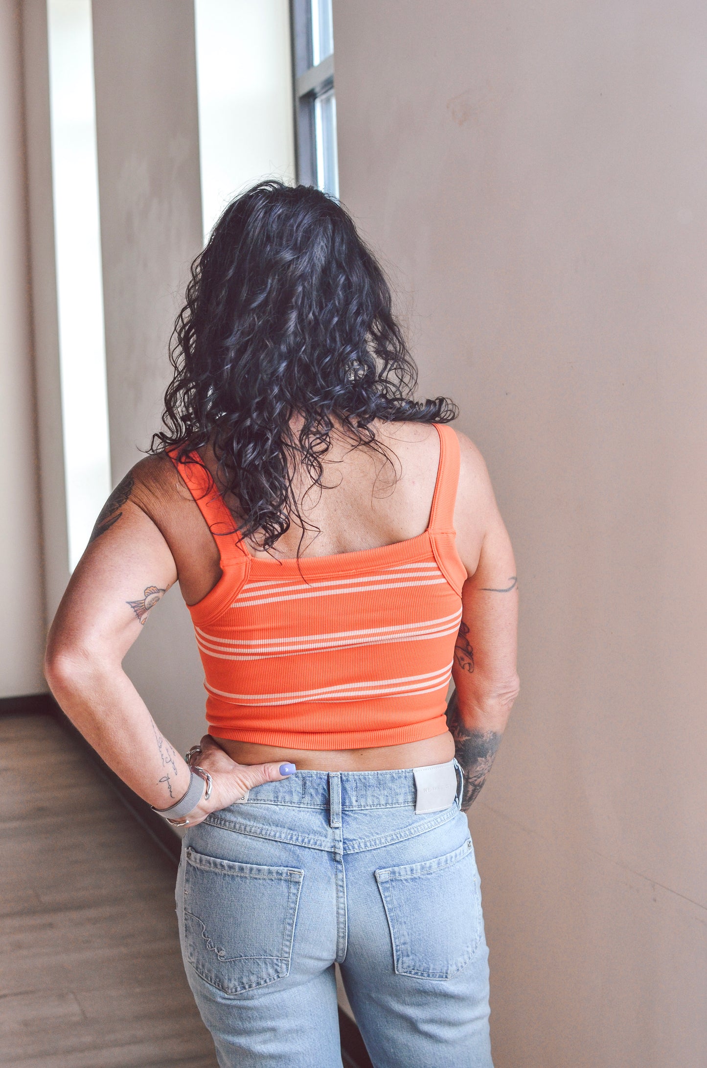 Person wearing an orange striped tank top and blue jeans standing against a neutral wall.
