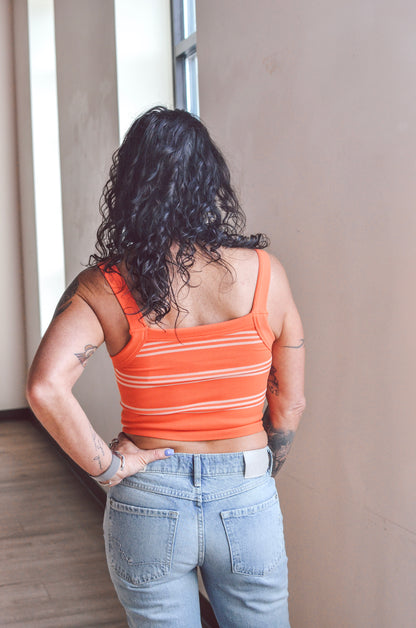 Person wearing an orange striped tank top and blue jeans standing against a neutral wall.