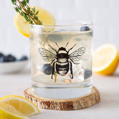 Glass with a bee design on a wooden coaster, surrounded by lemons and blueberries.