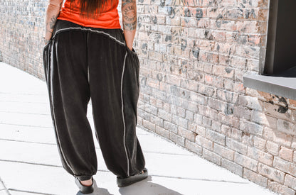 Person wearing black baggy pants with white piping standing on a rooftop with a brick wall background.