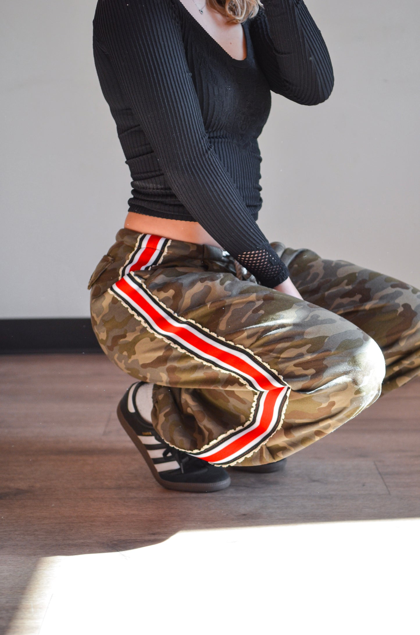 Person wearing camouflage pants with a red and white stripe sitting on a wooden floor.