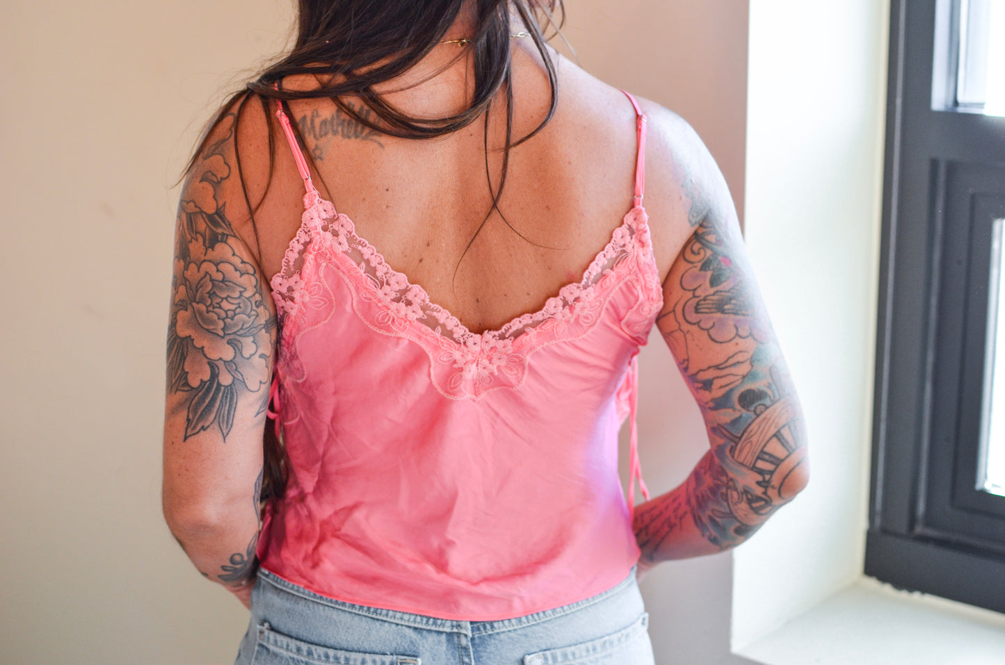 Person wearing a pink lace-trimmed camisole with tattoos on arms and back.