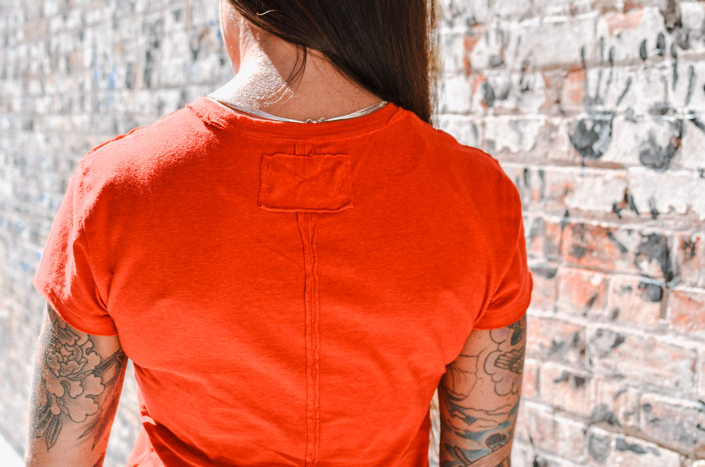 Person wearing an orange t-shirt with tattoos visible on arms, standing against a brick wall.