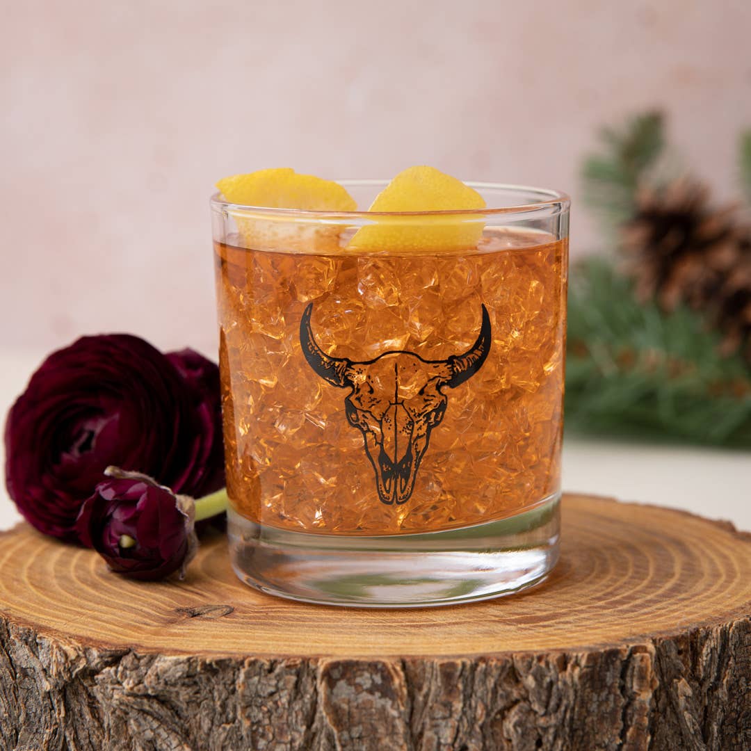 Glass of amber liquid with bull skull design on a wooden surface with flowers and pine cones.
