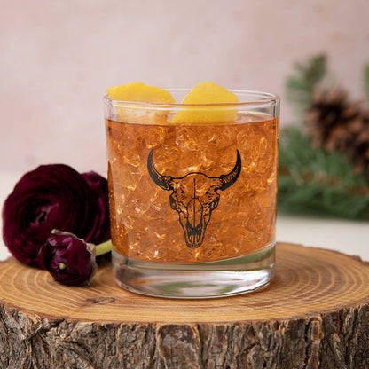 Glass of amber liquid with bull skull design on a wooden surface with flowers and pine cones.